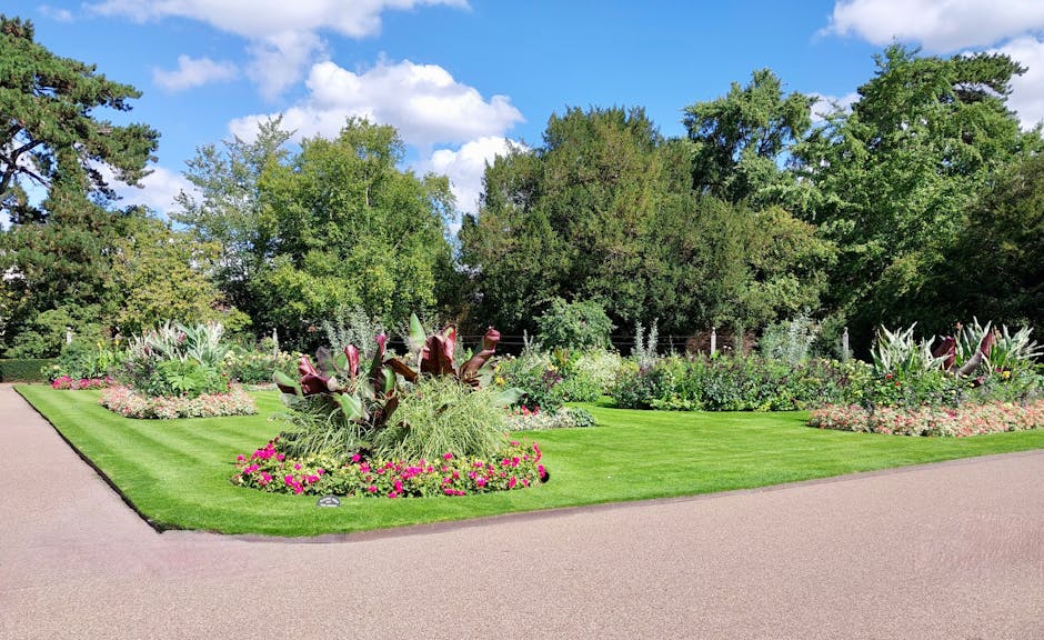 A well-maintained garden bed featuring a variety of ornamental plants and flowers, surrounded by neatly trimmed grass and laid out along a curved pathway. The bed includes large, dark-leafed plants wi